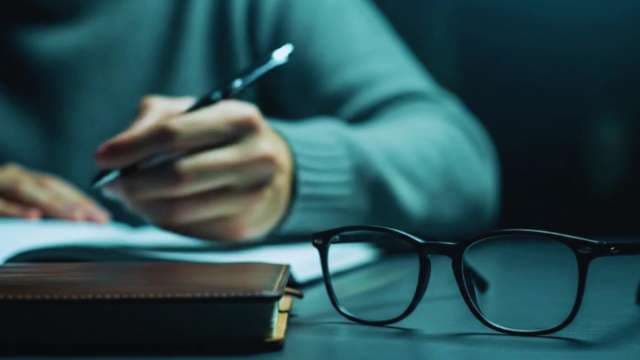 Person Writing with Pen and Eyeglasses in Moody Lighting