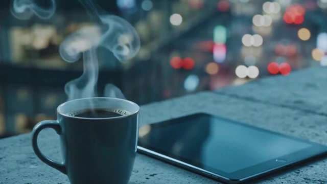 Steaming Coffee and Tablet Against Urban Night Cityscape