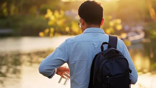 Man with Backpack Holding Water Bottle by Lake at Sunset