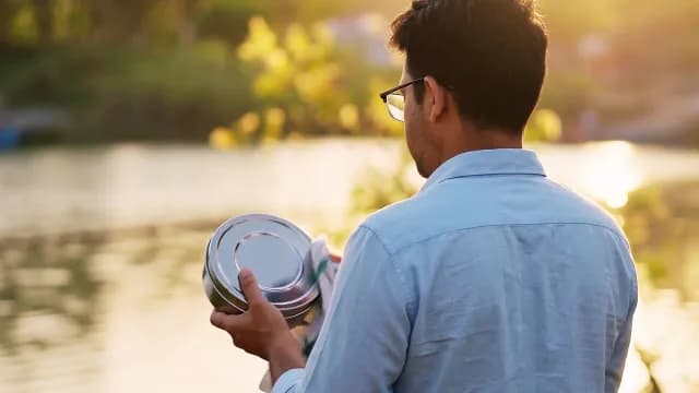 Man Holding Steel Lunch Box by Serene Lake at Sunset