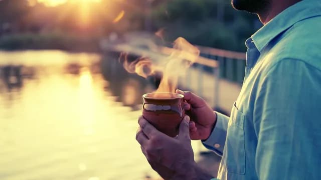 Man Holding Steaming Clay Mug at Golden Sunset by Lake