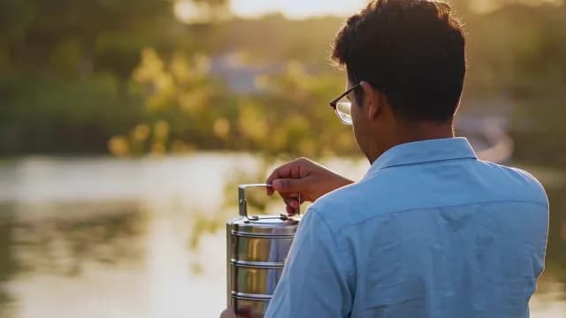 Man Holding Traditional Steel Tiffin Carrier by Scenic Lake During Sunset
