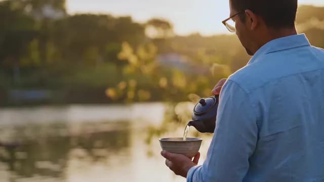 Man Pouring Hot Tea Into Ceramic Bowl by a Lake at Sunset