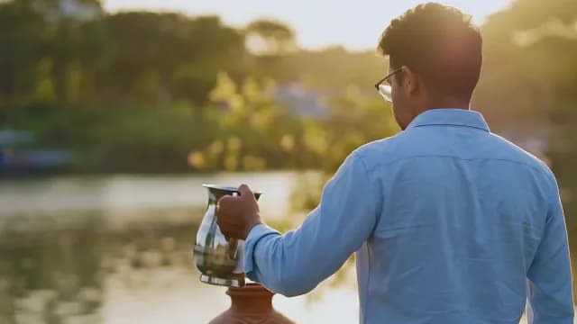 Young Man Pouring Water Into Clay Pot By Lake During Sunset
